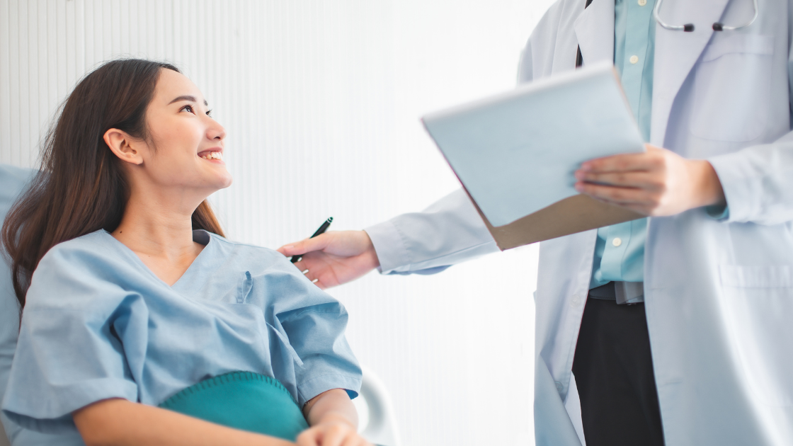 A smiling woman sits on an examination table with a friendly doctor standing beside her, preparing for a Pap test.