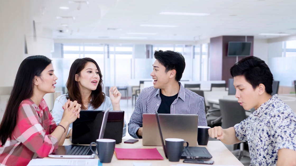 Four young Asian executives discussing work at an office desk, symbolizing corporate wellness and health screening SG