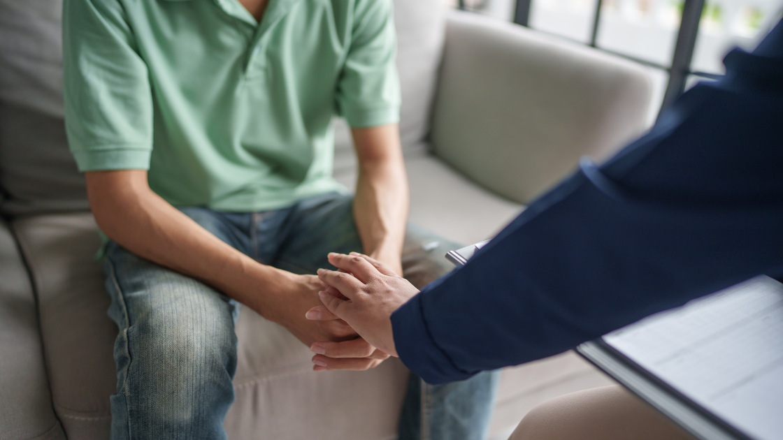 A man sitting with his head down while another person holds his hand in a comforting gesture, symbolising emotional support.