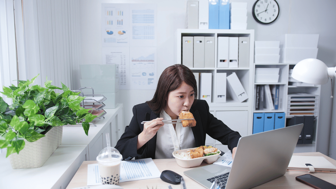 Asian female corporate worker eating fried chicken fillet with rice bento and boba milk tea at office desk showing common workplace nutrition choices