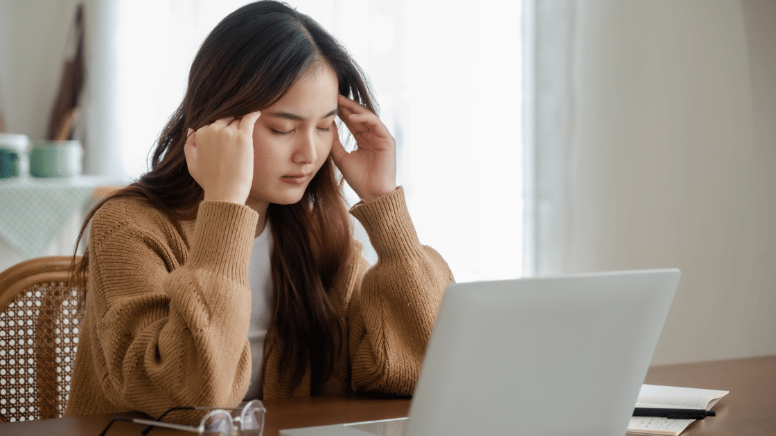 Stressed Asian female executive holding her head while sitting at office desk, representing workplace mental health challenges affecting corporate employees in Singapore
