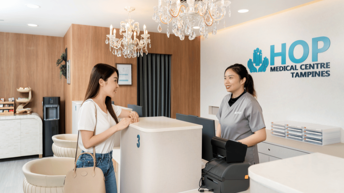 Friendly staff nurse at HOP Medical Centre reception counter warmly greeting female patient in modern Singapore health screening facility