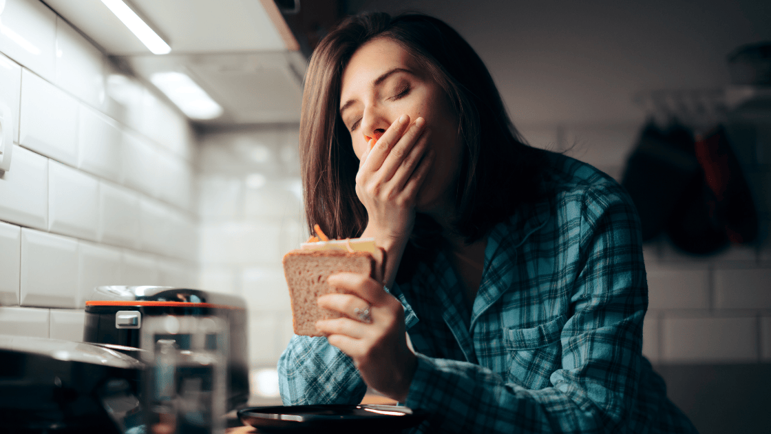 Tired woman yawning while holding bread and coffee, illustrating persistent fatigue from low cortisol symptoms